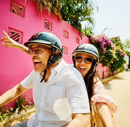 Couple in their 40's riding a scooter through a colorful neighborhood on a sunny day