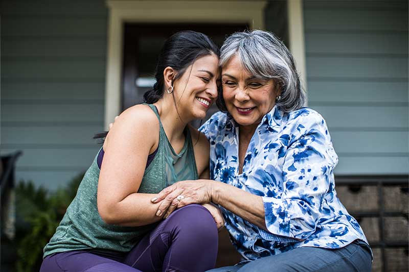 adult child and middle-aged parent sit closely and hold hands on front porch