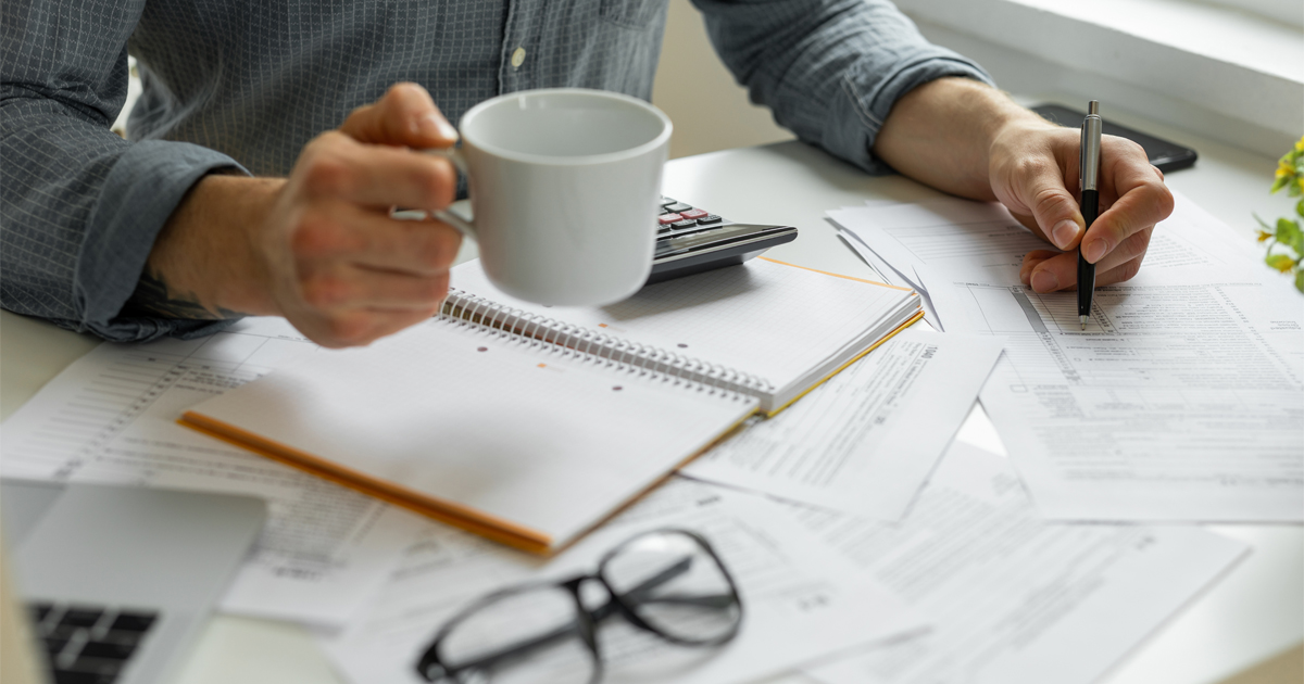 A person holds a mug in one hand and a pen in the other hand while doing paperwork.