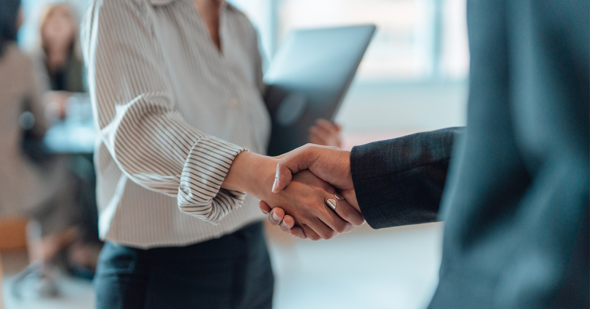 A man and woman shaking hands in a business setting. The woman is holding a laptop.