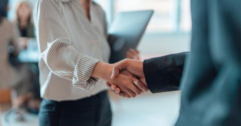 A man and woman shaking hands in a business setting. The woman is holding a laptop.