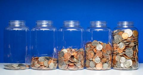 Five identical glass jars in front of a blue background each contain an increasing number of coins from left to right. The left-most jar only has a few coins while the right-most jar is nearly full of coins.